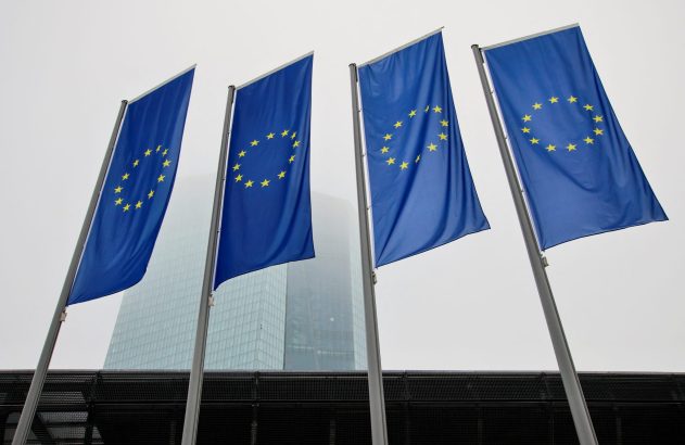 Quatro bandeiras da União Europeia exibidas em mastros altos. Ao fundo, um moderno arranha-céu de vidro sob céu cinzento.