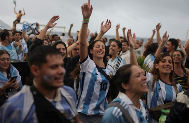Torcedores com camisas da Argentina celebram intensamente, levantando as mãos e sorrindo.