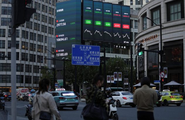 Tela exibe índices financeiros em rua de cidade chinesa com tráfego e pedestres.