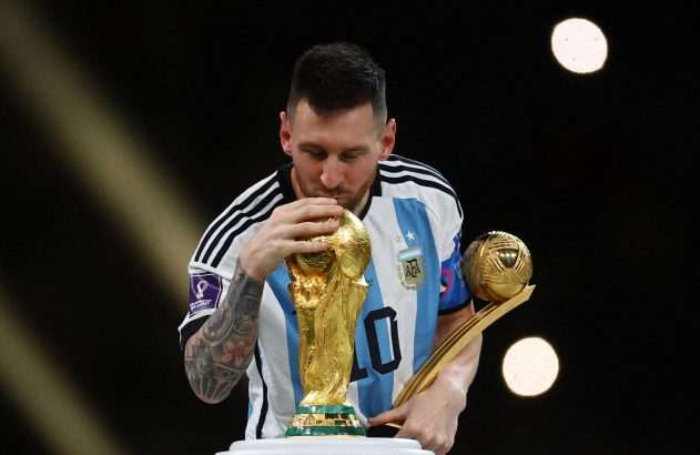 Lionel Messi, vestindo a camisa da Argentina, beija a taça dourada da Copa do Mundo enquanto segura outro troféu.