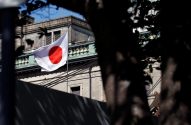 Bandeira do Japão, branca com círculo vermelho, tremula em frente a um prédio. Folhagem escura em primeiro plano.