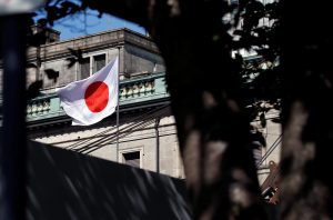 Bandeira do Japão, branca com círculo vermelho, tremula em frente a um prédio. Folhagem escura em primeiro plano.