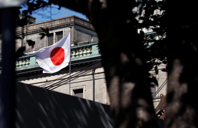 Bandeira do Japão, branca com círculo vermelho, tremula em frente a um prédio. Folhagem escura em primeiro plano.