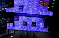 Tela azul de bolsa de valores com dados de empresas japonesas, observada por pessoas.