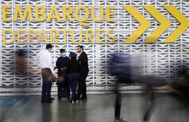 Pessoas em movimento em um saguão de aeroporto, com letreiro "EMBARQUE DEPARTURES" e setas amarelas.
