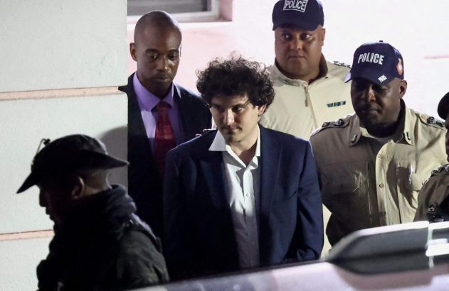 Sam Bankman-Fried é escoltado por policiais de uniforme e um homem de terno.