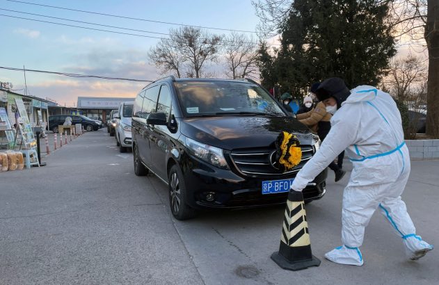 Pessoa em traje de proteção move cone ao lado de van preta com laço amarelo. Carros em fila ao fundo.