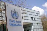 Placa branca da Organização Mundial da Saúde (OMS) com seu logo azul, em frente a um prédio moderno.