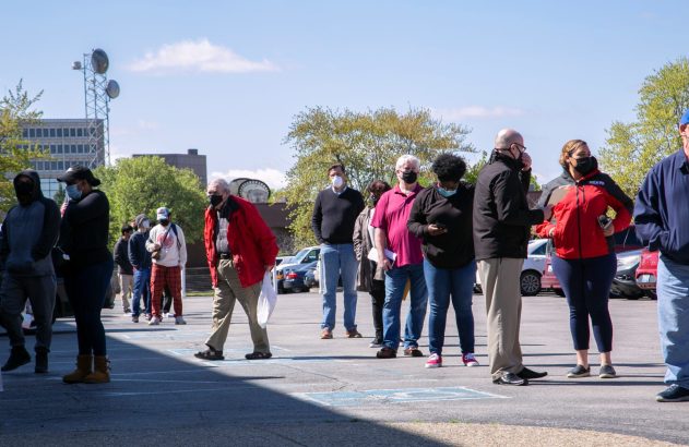 Fila de pessoas de máscara mantendo distanciamento em estacionamento com marcações no chão.