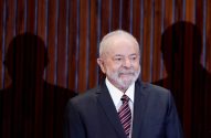 Luiz Inácio Lula da Silva, de barba branca, sorri em terno escuro. Sombras de pessoas no fundo.