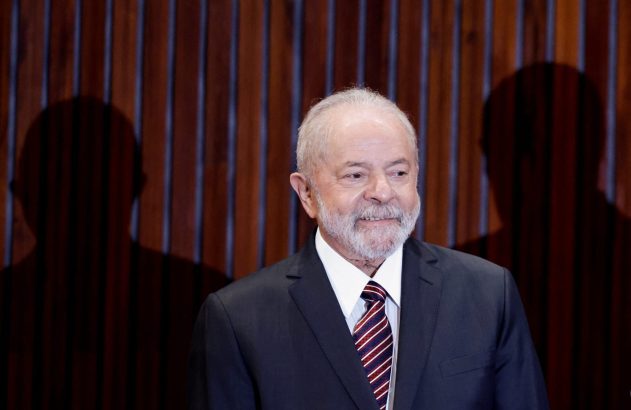 Luiz Inácio Lula da Silva, de barba branca, sorri em terno escuro. Sombras de pessoas no fundo.