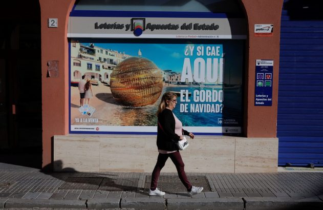 Mulher caminha em calçada ensolarada em frente à loja Loterías y Apuestas del Estado com anúncio do "El Gordo".
