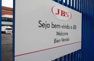 Placa JBS com 'Bem-vindo' em português, inglês e espanhol, e caminhão branco no pátio industrial.