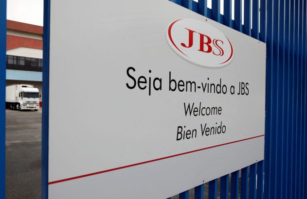 Placa JBS com 'Bem-vindo' em português, inglês e espanhol, e caminhão branco no pátio industrial.