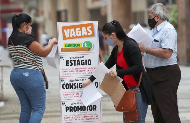Três pessoas, com máscaras, consultam um painel de vagas de emprego e manuseiam currículos.
