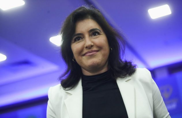 Mulher de cabelo escuro e blazer branco sorri levemente, olhando para cima, contra um fundo azul iluminado.
