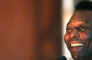 Pelé, em close-up, sorri mostrando dentes brancos e rosto iluminado. Fundo marrom desfocado.