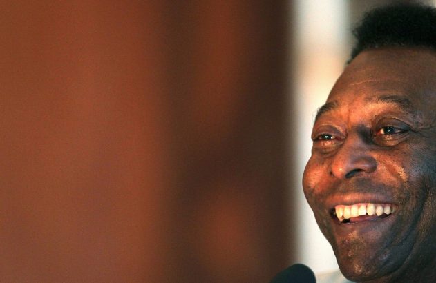 Pelé, em close-up, sorri mostrando dentes brancos e rosto iluminado. Fundo marrom desfocado.