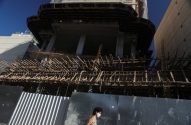 Prédio em construção com andaimes e fôrmas de madeira. Uma mulher de máscara caminha em frente ao tapume.