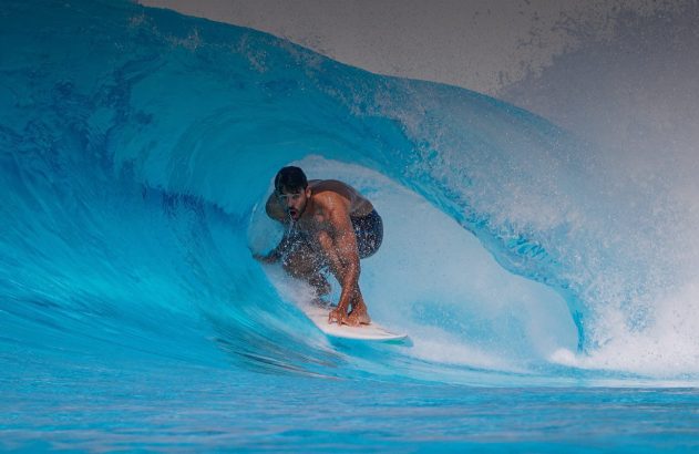Um homem surfando dentro de um tubo de uma onda azul grande. Muita água espirra em volta.