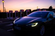 Carro Tesla preto em estação Supercharger ao anoitecer. Faróis acesos e carregadores iluminados sob céu colorido.