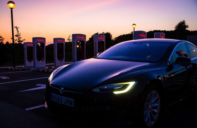 Carro Tesla preto em estação Supercharger ao anoitecer. Faróis acesos e carregadores iluminados sob céu colorido.