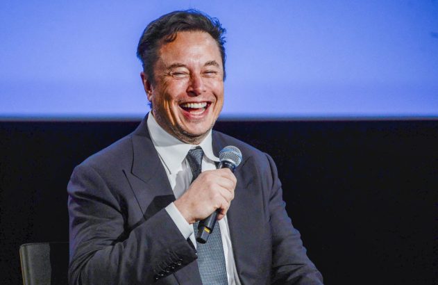 Elon Musk rindo muito, segurando um microfone, em um evento com fundo azul.