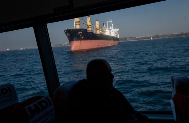 Vista de dentro de uma embarcação: homem em silhueta observa um navio de carga "MOHAMAD M" no mar.