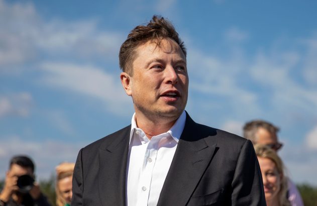 Elon Musk, de terno e camisa branca, olha para a direita. Ao ar livre, sob céu azul e pessoas borradas ao fundo.