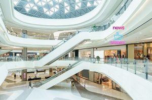 Interior de shopping moderno de múltiplos andares com lojas, escadas rolantes e teto de vidro. Pessoas circulam.