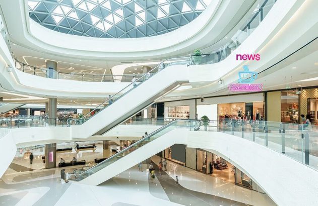 Interior de shopping moderno de múltiplos andares com lojas, escadas rolantes e teto de vidro. Pessoas circulam.