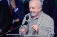 Lula sorri ao microfone em um pódio, usando blazer xadrez cinza e camisa cinza.