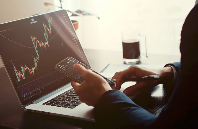 A imagem mostra uma os braços de uma pessoa que está mexendo no computador e no celular ao mesmo tempo. A pessoa analisa gráficos de variação de preços como exemplo de análises do mercado futuro.
