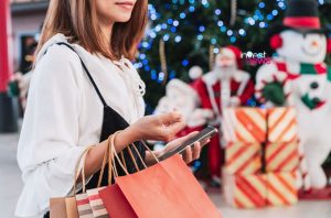 Mulher com sacolas de compras e celular, em ambiente natalino com árvore iluminada e decorações.