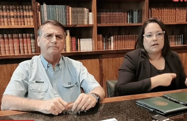 Jair Bolsonaro e mulher sentados em mesa. Ela gesticula, ele olha. Fundo com estantes de livros.