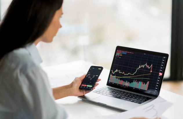 A imagem mostra uma mulher branca de cabelo liso e preto sentada frente a uma mesa branca. Ela segura um celular na mão e olha para a tela de um notebook. Os dois aparelhos mostram gráficos de variações no preço de uma ação.