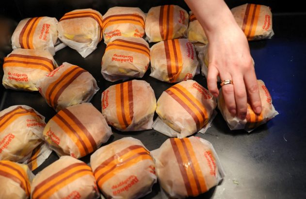 Mão alcançando um duplo cheeseburger embrulhado em papel listrado laranja e marrom, entre muitos outros.