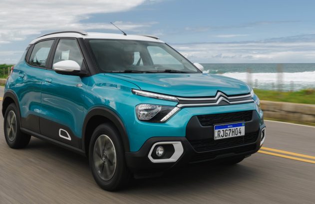 Citroën C3 azul-turquesa com teto branco dirigindo em estrada costeira, oceano ao fundo.