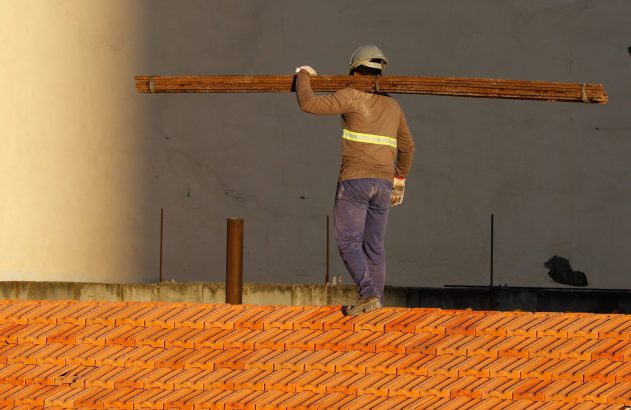 Trabalhador de capacete carrega vergalhões no ombro, em telhado de telhas.