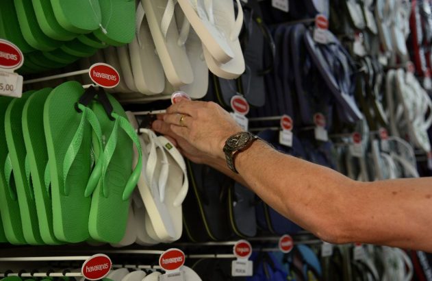Pessoa escolhendo chinelos Havaianas verdes em loja, com preço R$ 25,00.