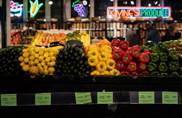 Mercado Lovine's Produce: bancas com abóboras, pimentões e pepinos frescos. Placas de preço e clientes.