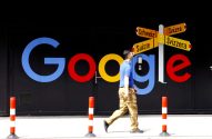 Homem caminha por fachada com logo colorido do Google. Sinal aponta Suíça em 4 idiomas. Postes de segurança.