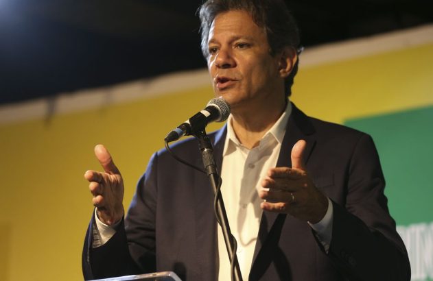 Homem em terno e camisa branca fala ao microfone, gesticulando. Fundo amarelo e verde.