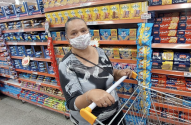Mulher de máscara facial empurrando um carrinho de compras em corredor de supermercado com prateleiras cheias.