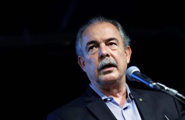 Homem de bigode e cabelo grisalho, de blazer, falando ao microfone com fundo escuro. Olhando para cima.