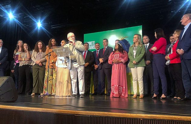 Presidente Lula discursando em palco, ladeado por autoridades e uma mulher indígena, em evento oficial.