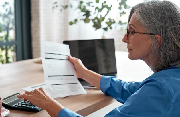 A imagem mostra uma mulher idosa sentada em frente a uma mesa de madeira. Ela segura um documento com dados em uma mão, enquanto faz contas em uma calculadora. (Foto: Shutterstock / Ground Picture)
