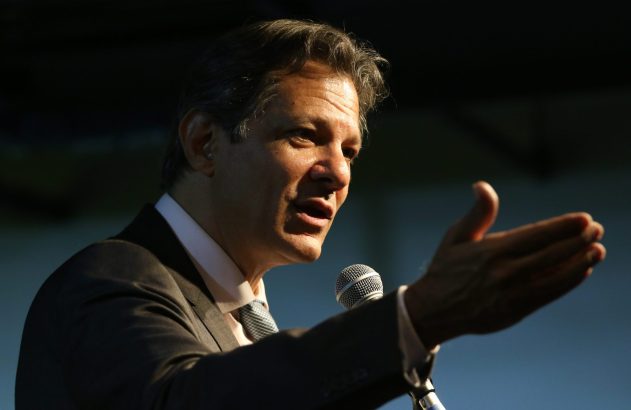 Fernando Haddad discursando em um microfone, com uma das mãos estendida.