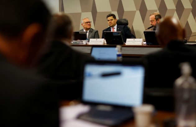 Três homens em uma mesa de audiência. Dois senadores à frente, um à direita. Laptops e participantes em primeiro plano.