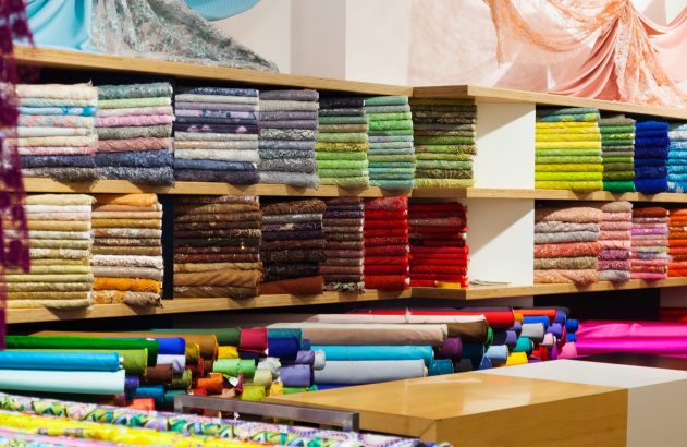 Loja de tecidos com prateleiras repletas de pilhas e rolos de tecidos coloridos e texturizados, alguns expostos no alto.